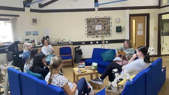 A group of women sit on blue sofas arranged in a square, looking at one woman playing the guitar.