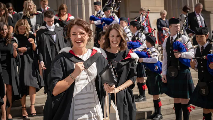 Two girls walking down steps outside of a building, wearing graduation robes. A group of graduates stand behind them, and a row of bagpipers stand lined up on the steps.