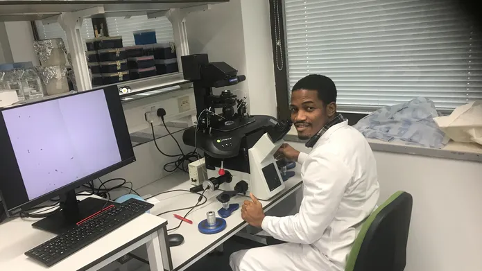 A man wearing a white lab coat sits at a desk with a computer and other gadgets.