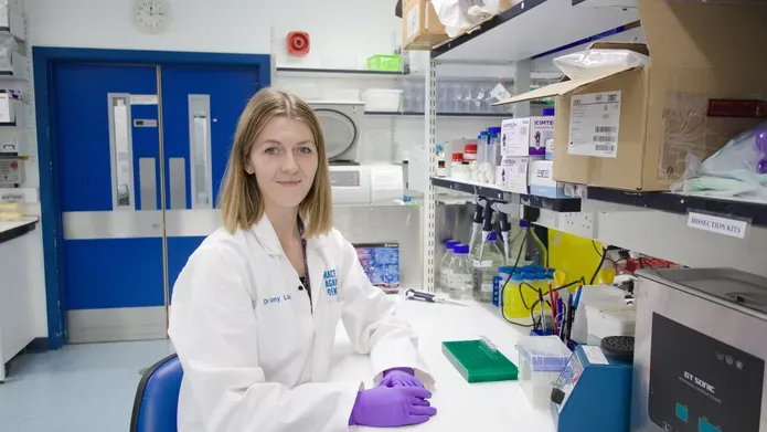 Woman wearing white lab coat, sat at a table wearing purple gloves, in front of shelves containing science equipment