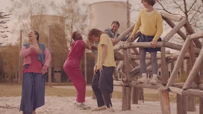 Film still. Six dancers in brighly coloured clothing hang out in a public park