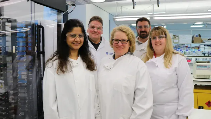 Group of scientists wearing lab coats and protective eyewear, three woman in front and two men in row behind, standing in a lab with science equipment around them. 