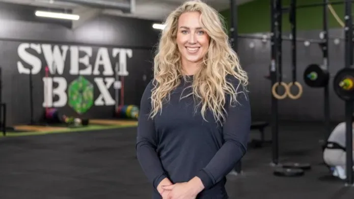 woman with long, wavy blond hair looking at camera wearing a black long sleeved top, with gym equipment in the background