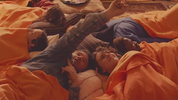 Film still. A group lie down wrapped in orange blankets, their heads almost touch forming a cirle
