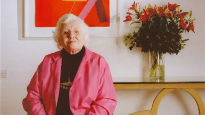 Wilhelmina Barns-Graham  sits in front of a red print and bunch of flowers, wearing a pink blazer.