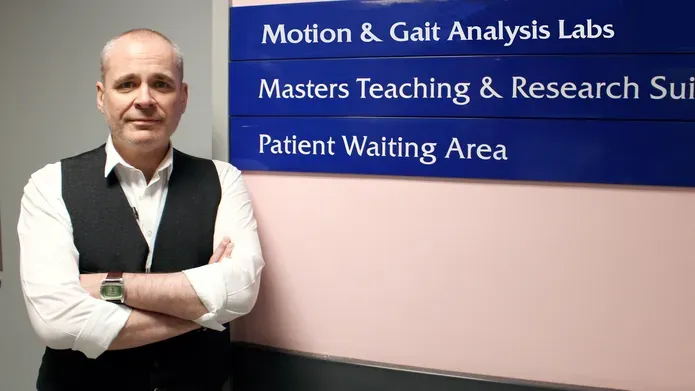 A man wearing a white shirt and black waistcoat stands next to a blue sign.