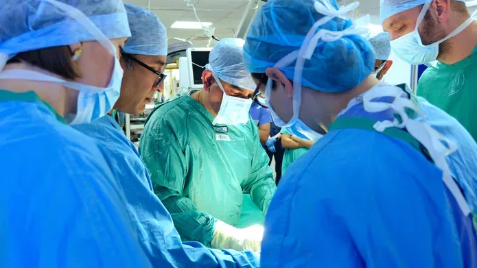 Surgeons wearing blue and green clothing, masks and hair nets, stand around looking at a demonstration together.