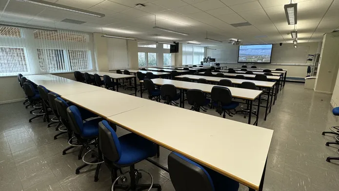 A room full of desks and blue chairs with wheels. There are windows along the left wall.