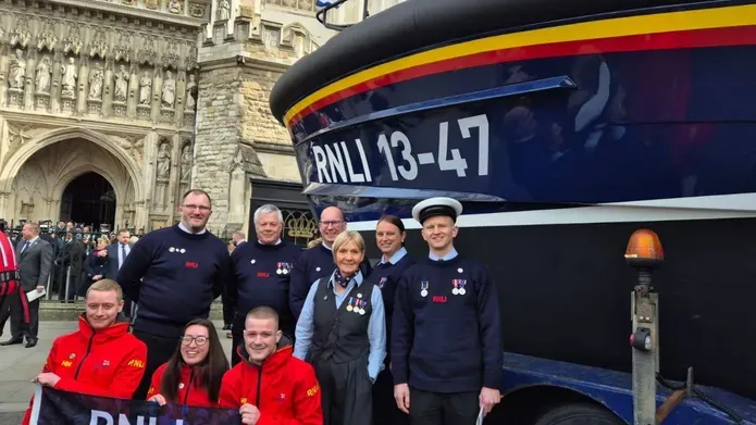 Lifeboat outside Westminster Abbey, with a group of people standing to one side of it. Front row of three people wearing red outfits hold a flag while six people behind wear dark uniforms and medals.