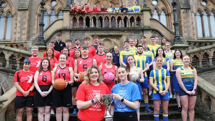 Large group of people wearing sports kit standing on outside steps, one half wearing red kits and the other half wearing blue and yellow striped kits. Front two women hold a trophy between the two of them.