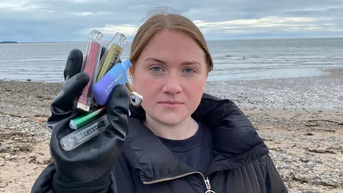 Woman on beach holding up a handful of discarded disposable vapes, wearing a big jacket and looking stern. 