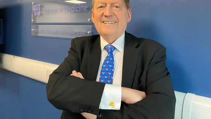 Professor Tom MacDonald stands with arms folded in front of a blue background.