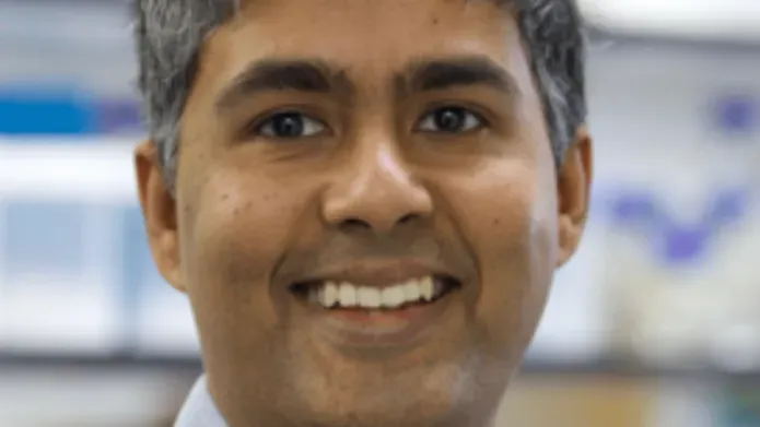 man in a blue shirt and grey jumper is stood in a lab smiling at the camera