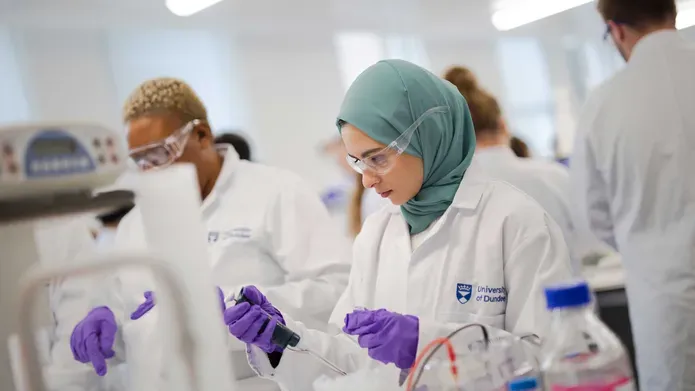Two females in lab coats, purple gloves and safety glasses. One girl is holding an instrument in her hand and looking at it.