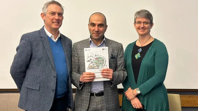 Three people grouped together. A male in the centre holds a booklet and there is a male on his left and a female on his right. All are smartly dressed and smiling