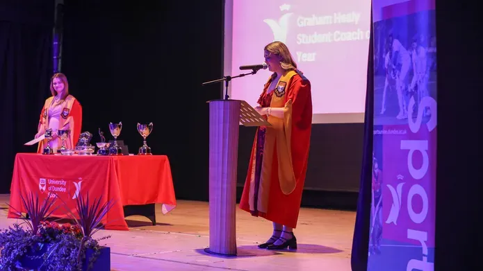 A women in a robe stands in front of a pedestal speaking in a microphone. Another person stands behind a table covered in red cloth.