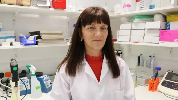 Woman with long dark hair, wearing white lab coat, standing in a lab with shelves behind her with lots of science materials on them.