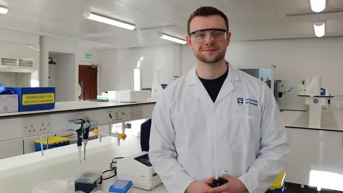 Young man wearing white lab coat and protective eye wear, standing in a science lab with science equipment around him