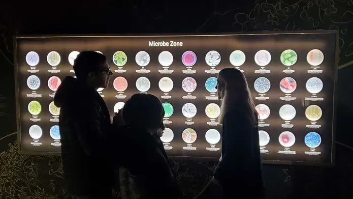 Professor Sarah Coulthurst speaking to members of the public in front of a wall displaying lots of microbes at Dundee Science Centre