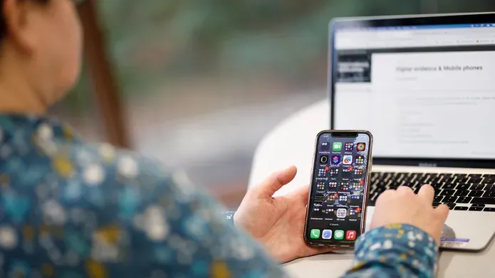 A person sitting at a table with a mobile phone in one hand and a laptop on the table
