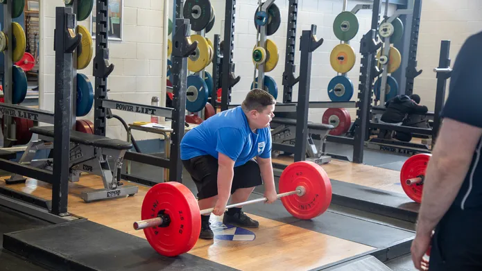 A boy squats over a set of weights, ready to lift. There are lots of weights stored on racks around the room behind him.