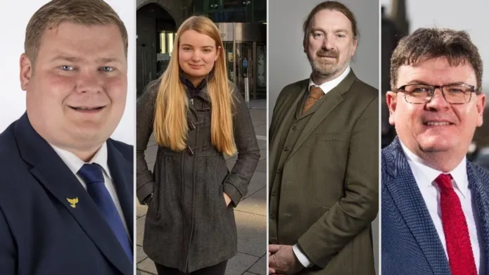 From left to right; a mean in a blue suit and blue tie with a white shirt smiling, a woman with long blonde hair wearing a grey jacket with her hands in her pockets, a man in a green tween suit with a yellow tie, a man with glasses wearing a blue suit with a red tie.