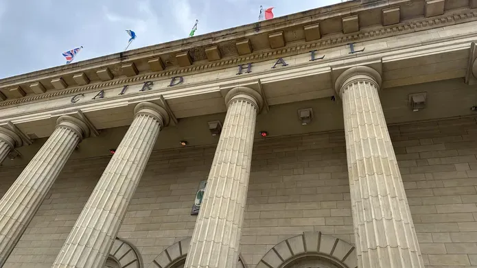 Roof area of old building with tall pillars and flags out of the top, and a cloudy blue sky above