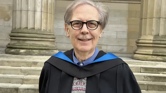 Man aged 75 with greying hair parted at the side and down to eye level, wearing glasses and graduation gown and hood and smiling at the camera