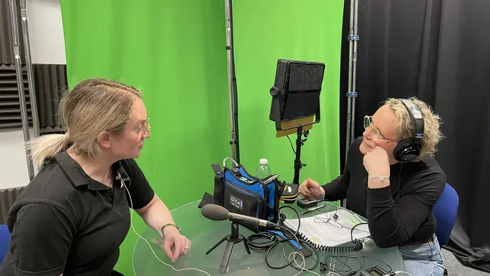 Two women sitting down at a table facing each other and talking, both wearing headphones. On the table are microphones pointing at each of them and lots of audio recording equipment.