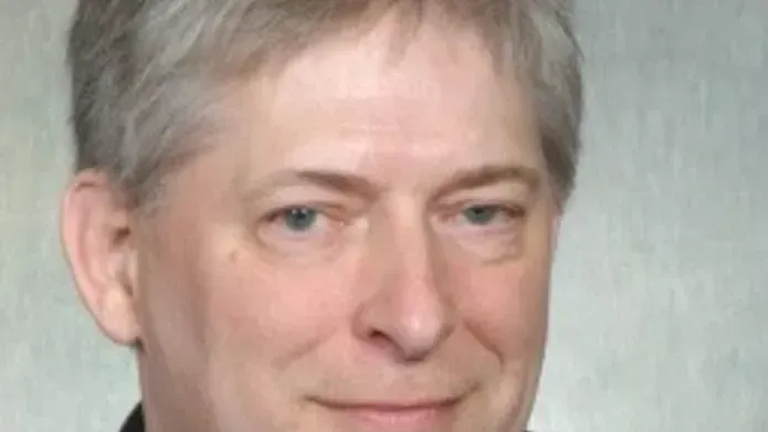 Man with grey hair in a shirt, tie and blazer against a grey back ground with a slight smile