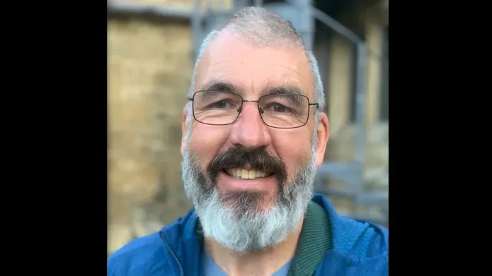 man in a blue shirt and grey jumper is stood in a lab smiling at the camera