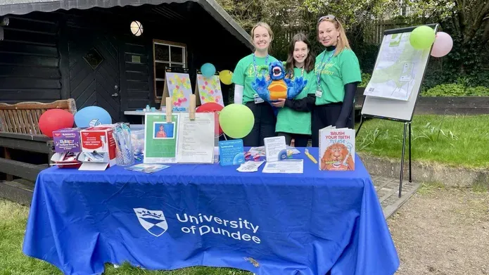 Three people stand in front of a table filled with colourful displays, with an outdoors garden in the background.