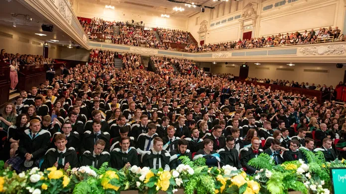 Hundreds of students sitting together in a large hall for their graduation ceremony. There is a upper level for their families.  