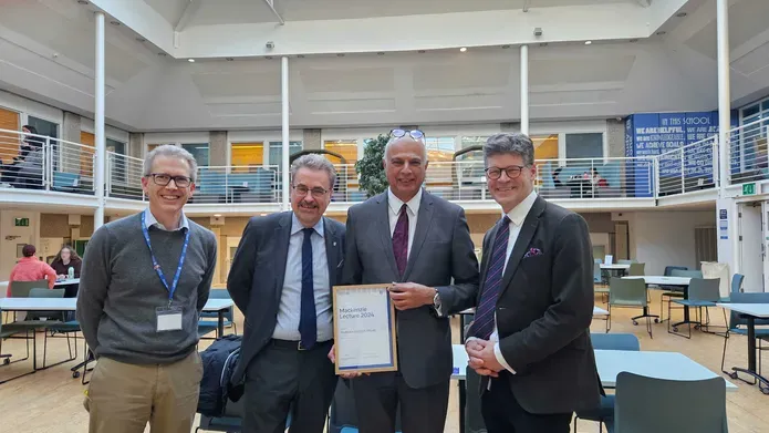 Four men wearing suits stand together holding a framed certificate for the Mackenzie Lecture 2024