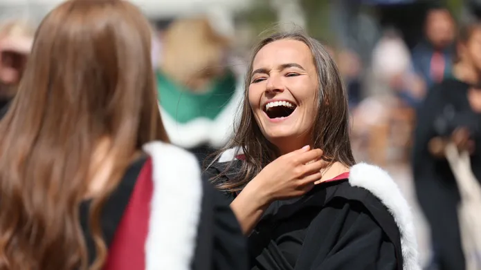 Two women in graduation gowns with red hoods lined with white fur. One stands with her back to us, the other faces us and is laughing.