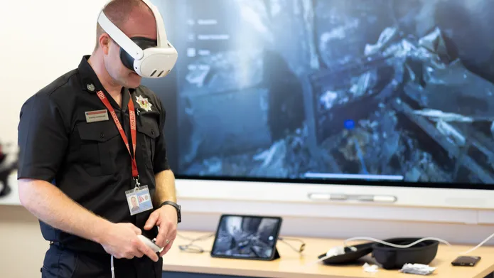 Man wearing fire fighter uniform and virtual reality headset over his eyes, with a large screen behind him showing a fire scene