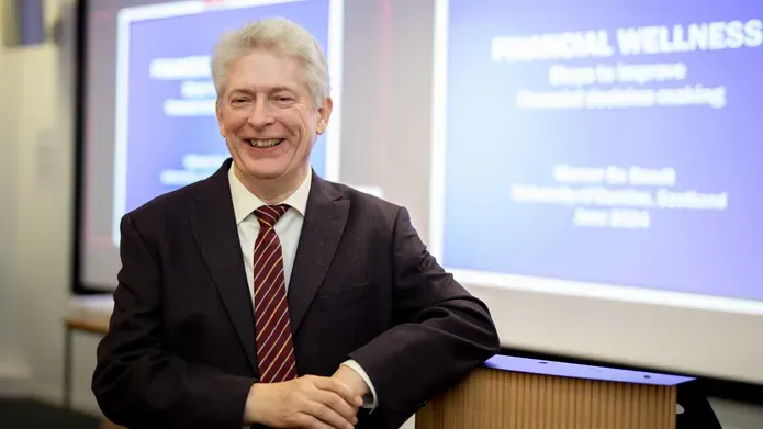A man wearing a suit with a red tie leans against a podium, there are screens behind him showing a presentation.