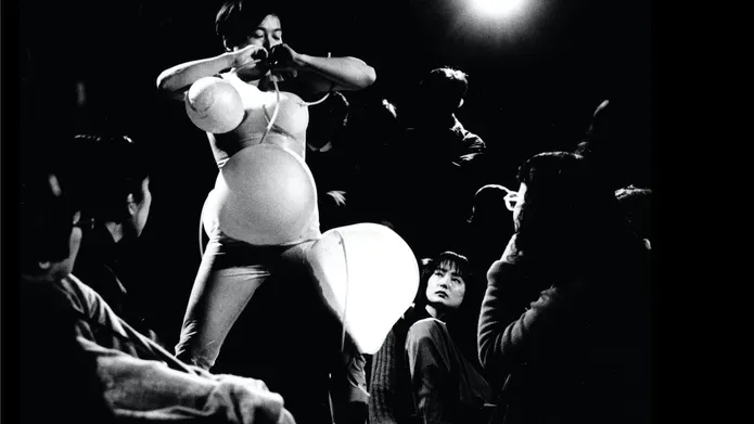 Tari Ito performance view black and white. Artist blowing inflatables around her body with tubes connected to her mouth creating enlarged spheres on her stomach, chest