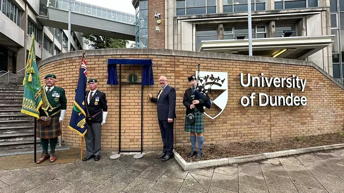 Two men in formal wear holding flags, one man in a suit standing next to a plaque and a piper