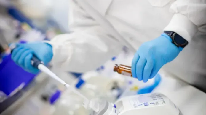 A person pipetting into a brown vial wearing a lab coat and blue gloves