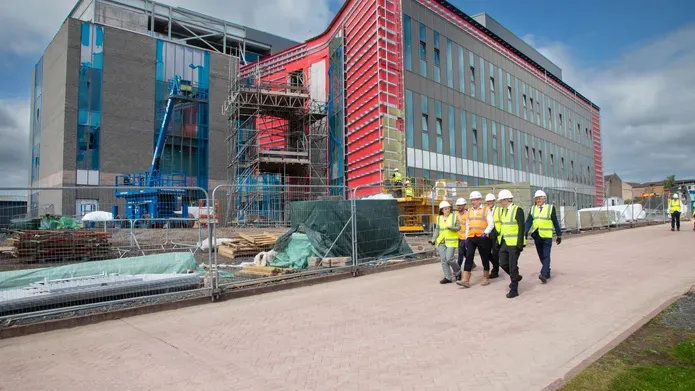 A group of people in high-vis vests walking in front of a building site. There is a very large building in the site.