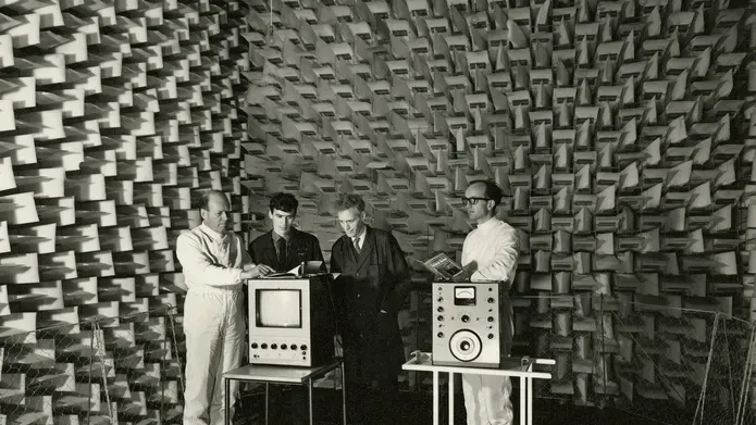 Four men stand inside an Anechoic Chamber. They are looking at two pieces of equipment, one a monitor, the other some sort of measurement device