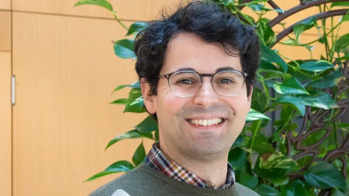 A man with short dark brown hair smiling. He is wearing glasses and a dark green jumper. Behind him are the leaves from a plant.