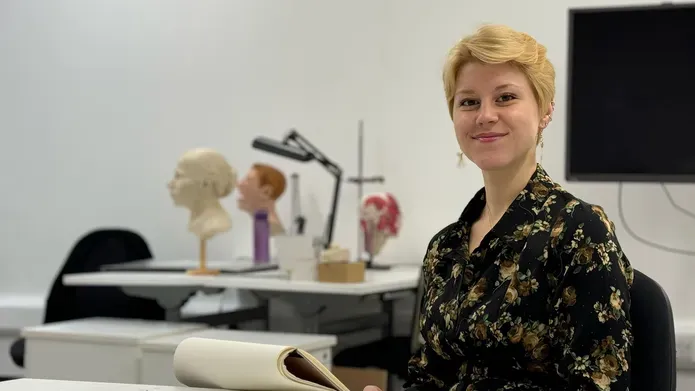 Cydney Davidson sits at a desk, smiling, with a sketch notebook placed in front of her