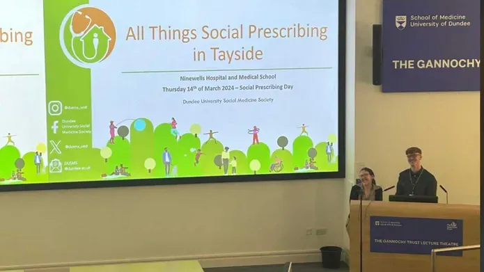 Two smiling university students stand at the front of a lecture theatre.
