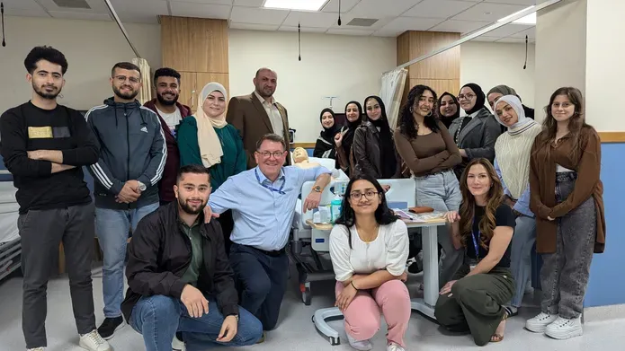 A group of visiting students pose around a bed with a member of staff from Dundee. While the bed appears to be a standard hospital ward bed, it in fact has a surgical dummy lying on it as this entire room is a simulation ward used for training.