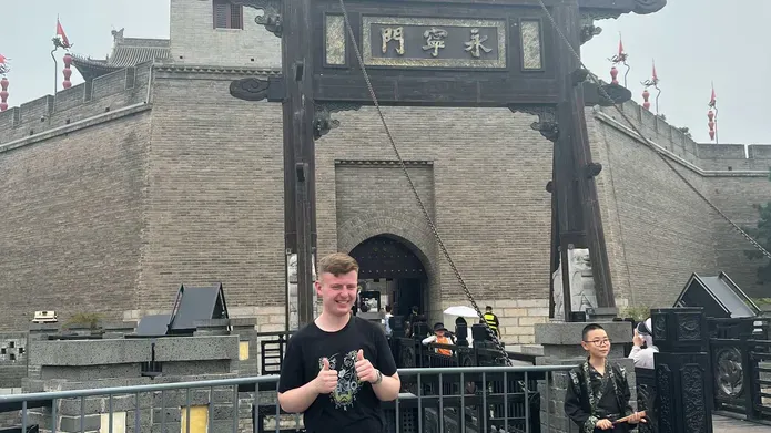A man in a black tshirt and blue jeans stands in front of a Chinese building with two thumbs up
