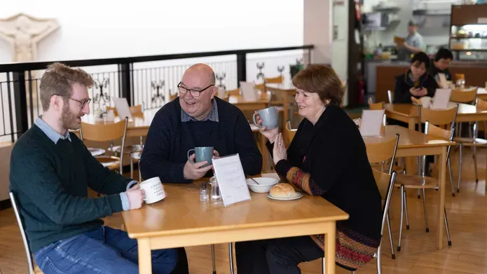 Three people sit around a wooden table smiling and drinking from mugs.