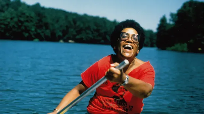 Photo of Audre Lorde on a lake laughing. She is a black woman and is paddling, laughing, wearing afro hair, glasses and a red t-shirt
