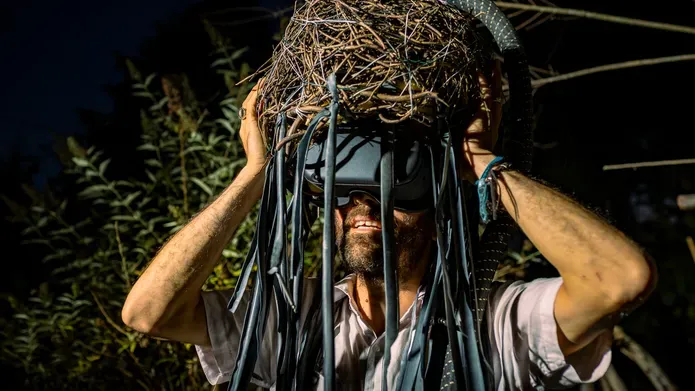Man with a VR head set with blue fabric and twigs hanging from his head